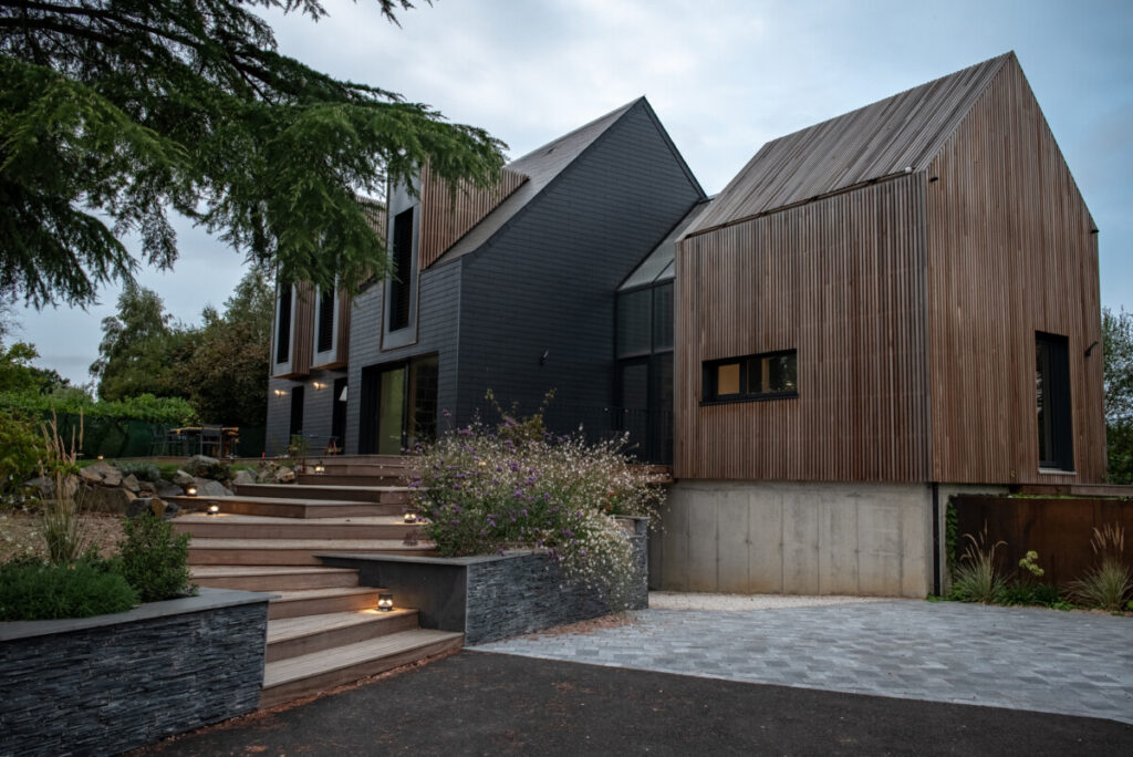 Vue sur la maison réhabilitée, bardage noir et extension en bois, verrière entre les deux pour marquer l'entrée.