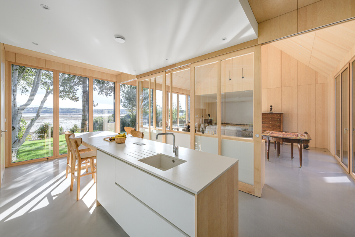 Intérieur de la maison passive, en bois clair et chaleureux, avec une grande baie vitrée offrant une belle vue sur la mer. Au premier plan, une cuisine avec plan de trvail sobre et blanc et une verrière intérieure en bois